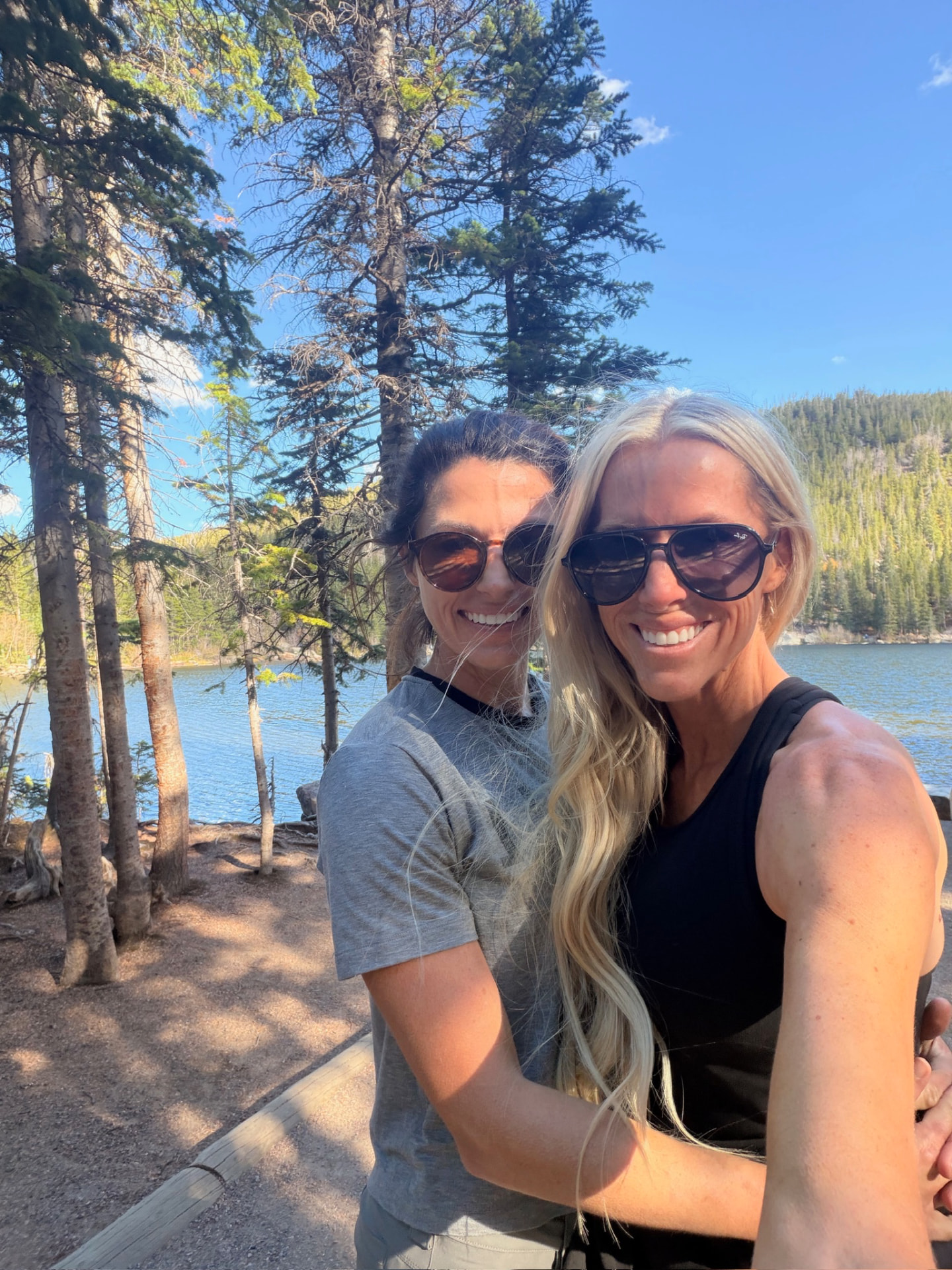 Mary and Sarah together beside an alpine lake, pine trees and a mountain slope behind them